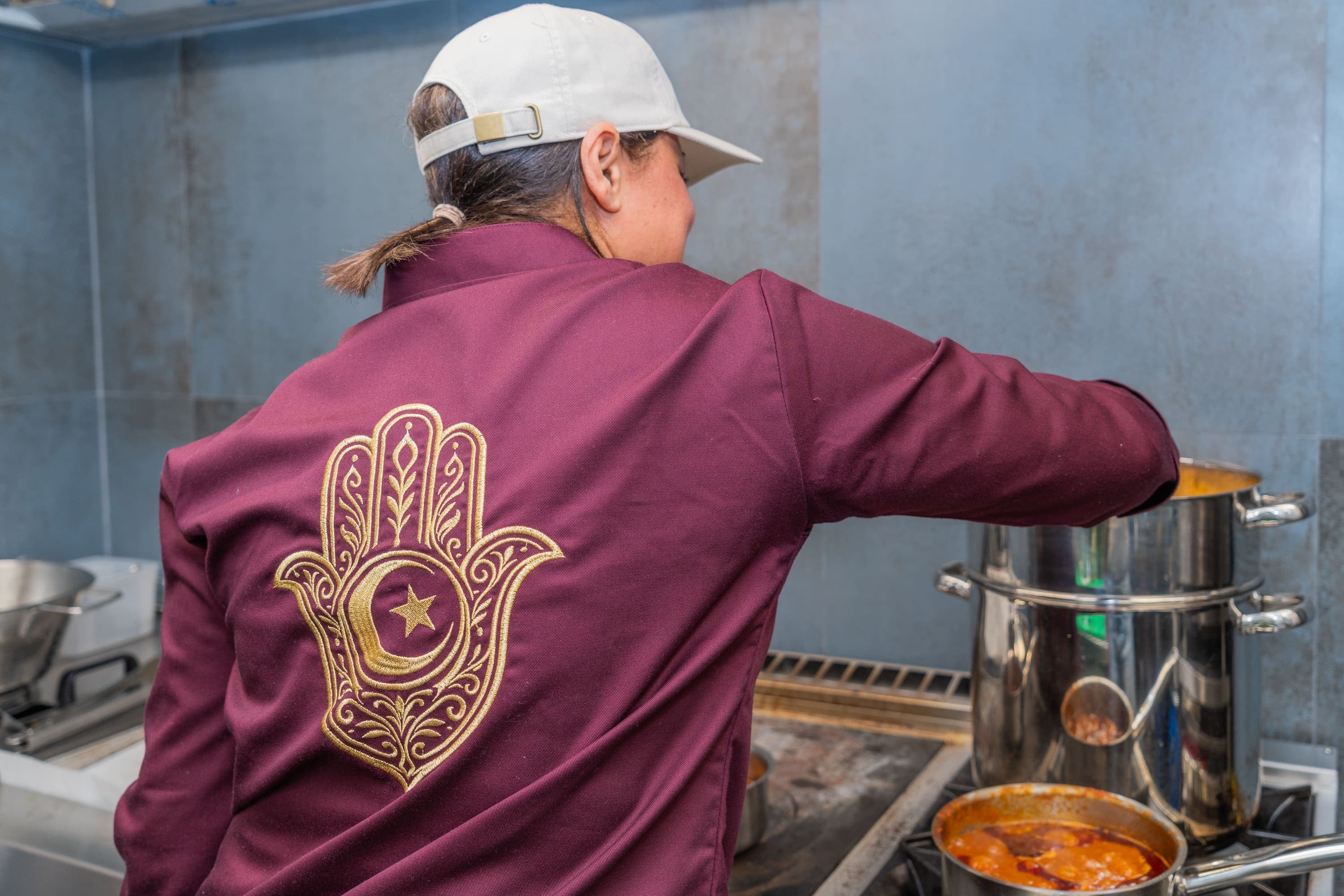 En cuisine, de dos, veste bordeaux brodée d’une main de Fatma dorée, devant les marmites.